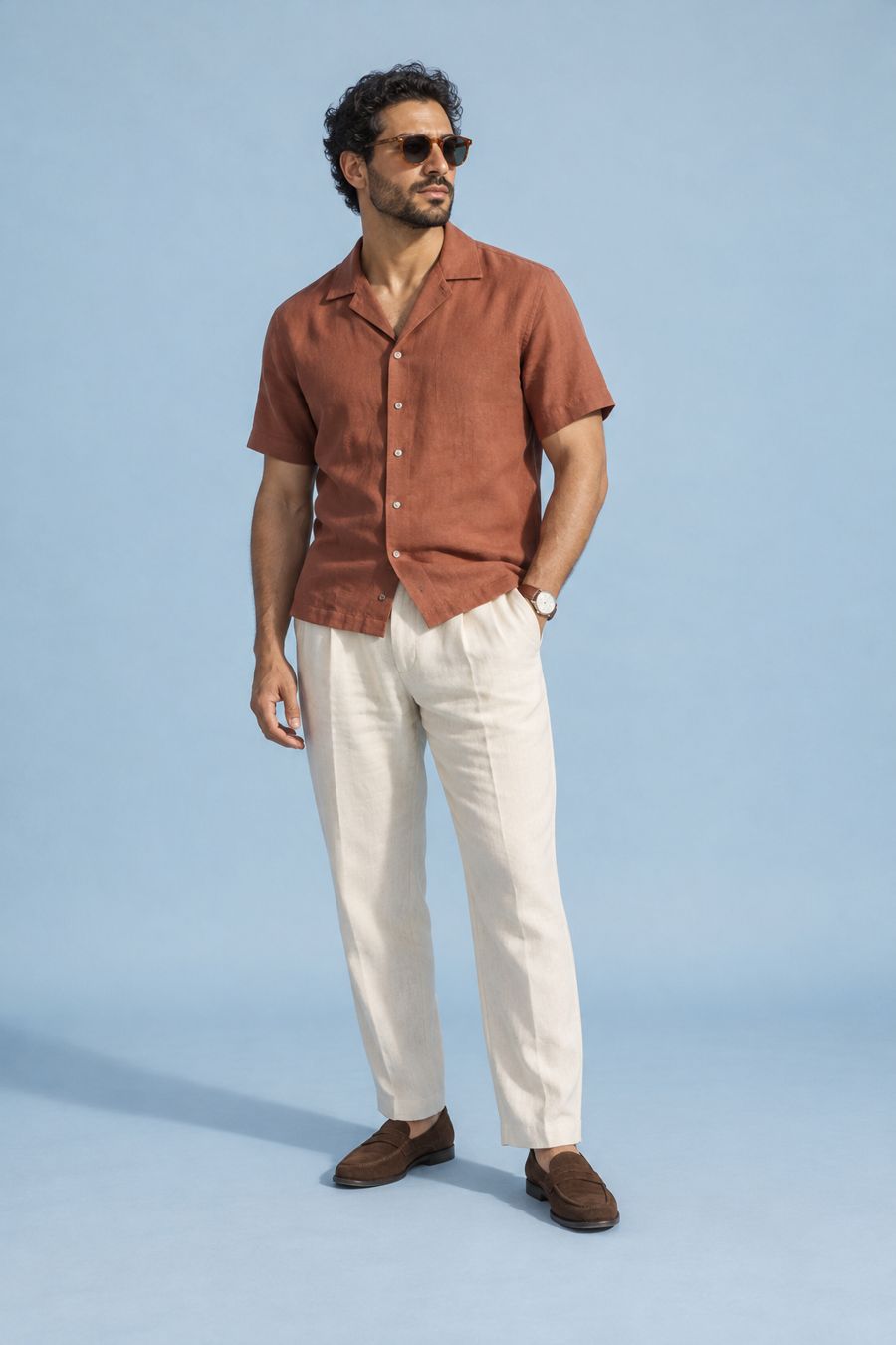 A man in a terracotta camp-collar shirt, cream linen trousers, brown suede loafers, sunglasses and a leather-strap watch.